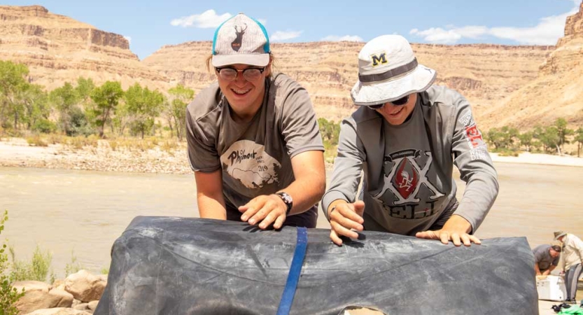 Two people roll up a large tarp. Behind them is a river and and very tall red canyon walls